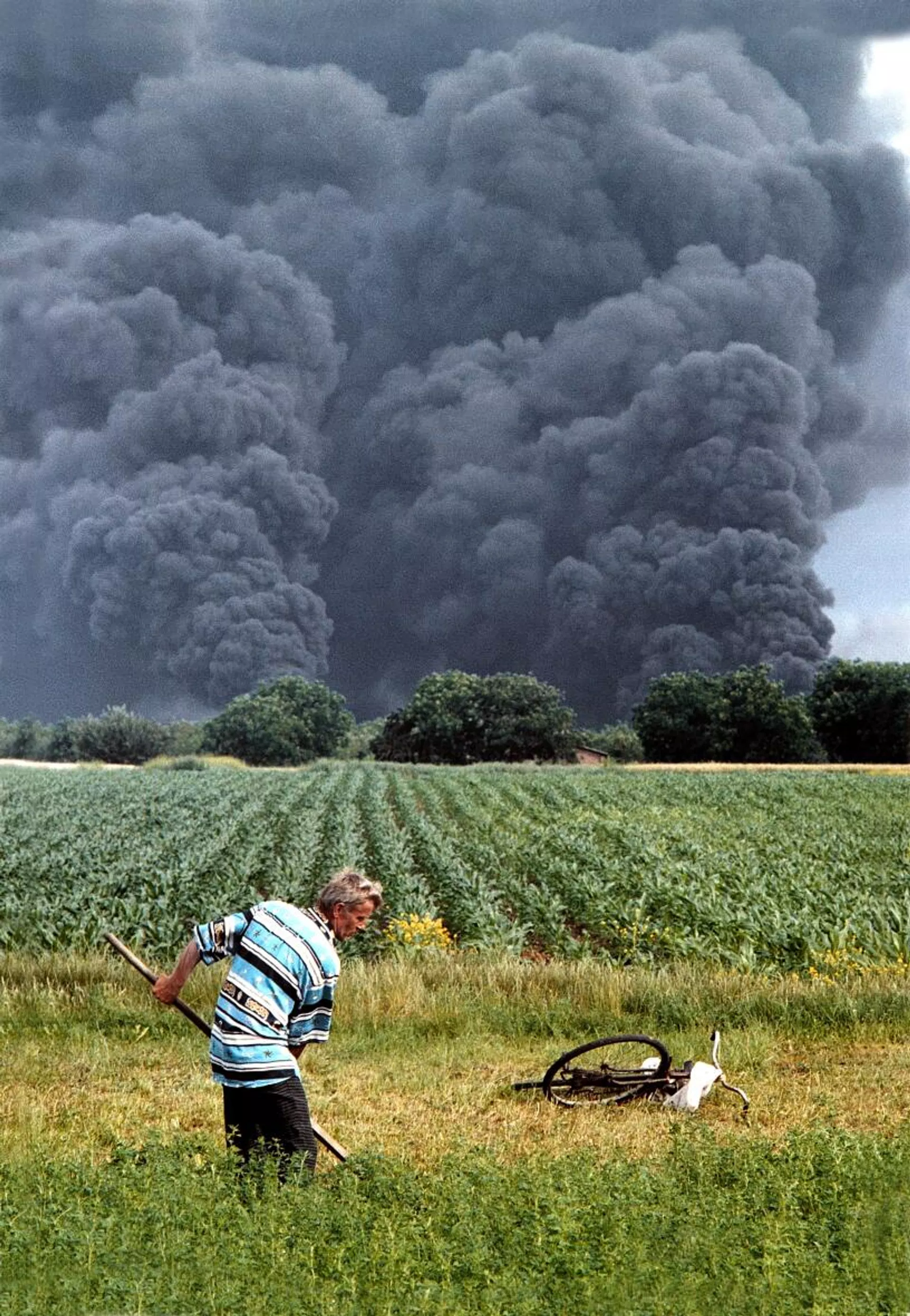 Photograph of a man mowing grass by hand in a green field, with a massive column of black smoke rising from a burning refinery in the distance behind him.